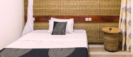A neatly made bed with a white and black bedspread, a woven headboard, and a small round side table with a remote control.
