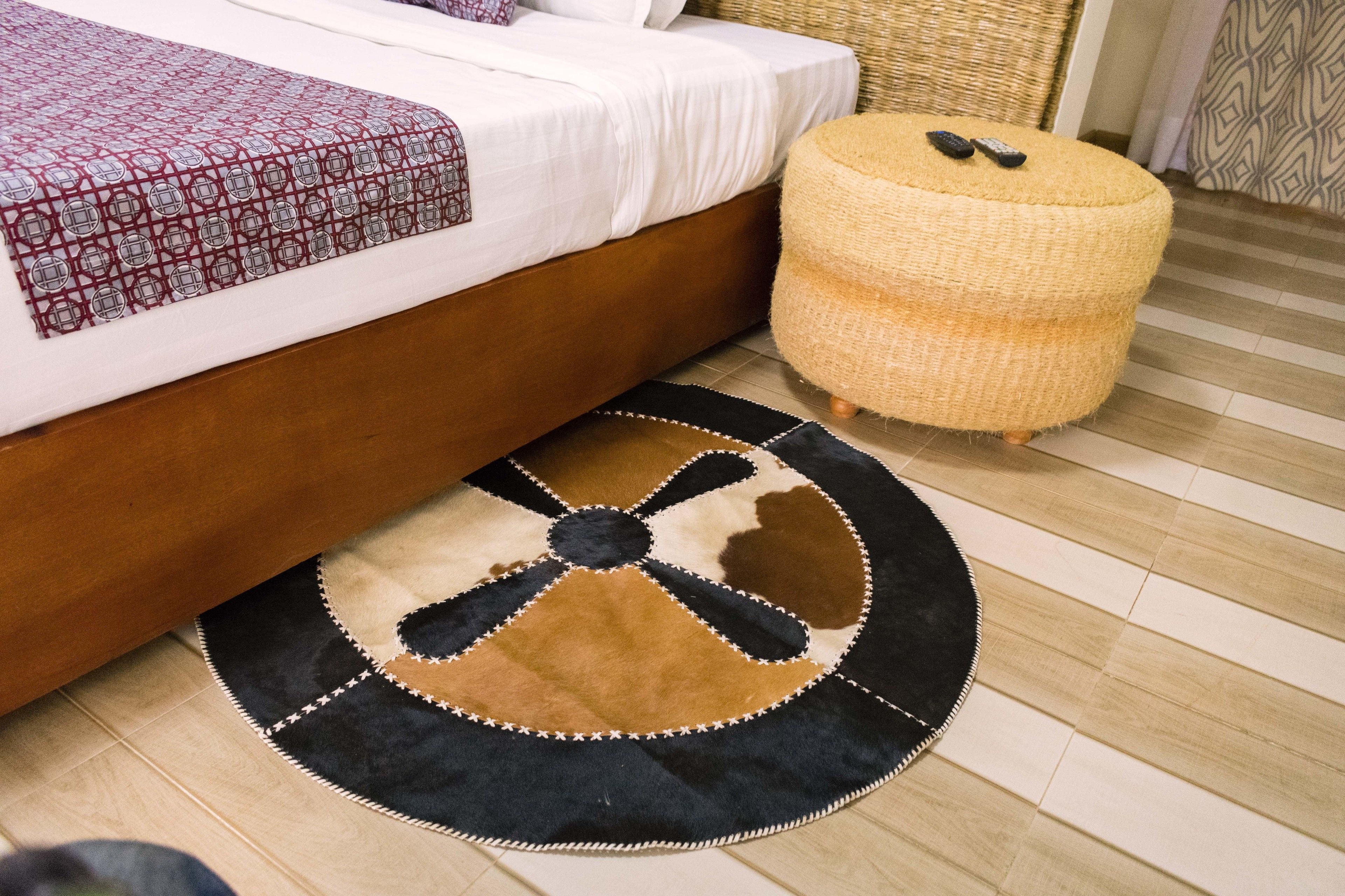 Een net opgemaakt bed met een bedsprei met patroon, een rond vloerkleed met een ontwerp in het midden, en een geweven pouf met afstandsbedieningen erop.