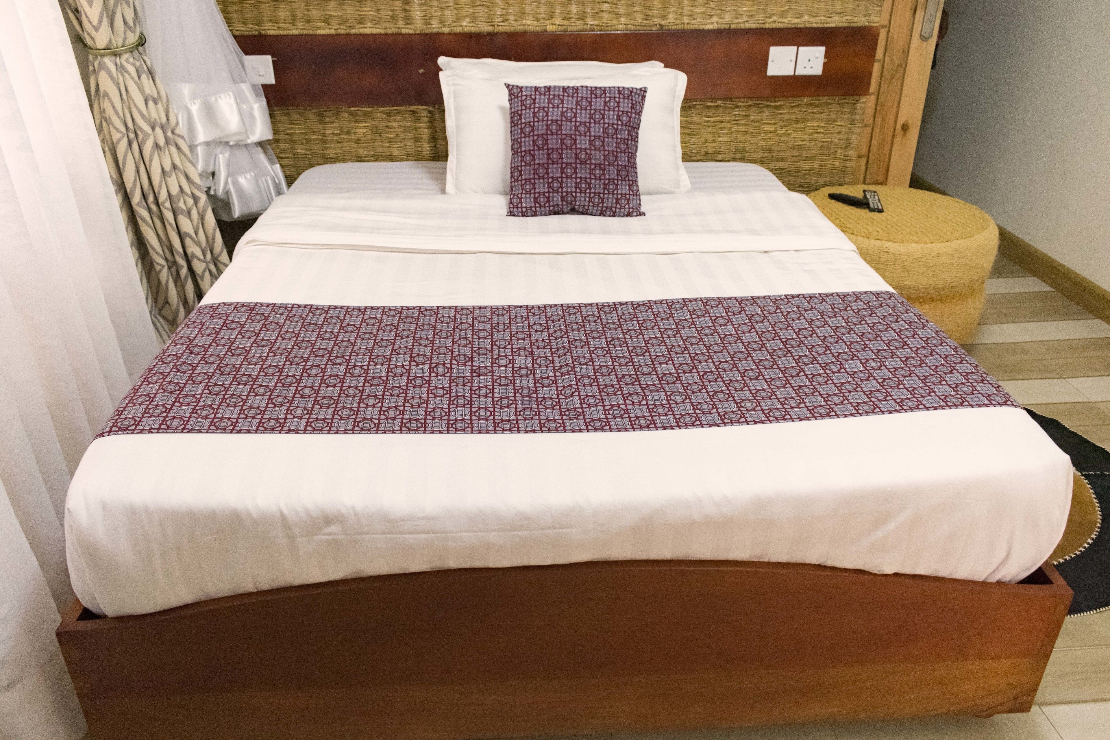 A neatly made bed with a patterned bedspread and pillows, a wooden headboard, and a small ottoman with a remote control.