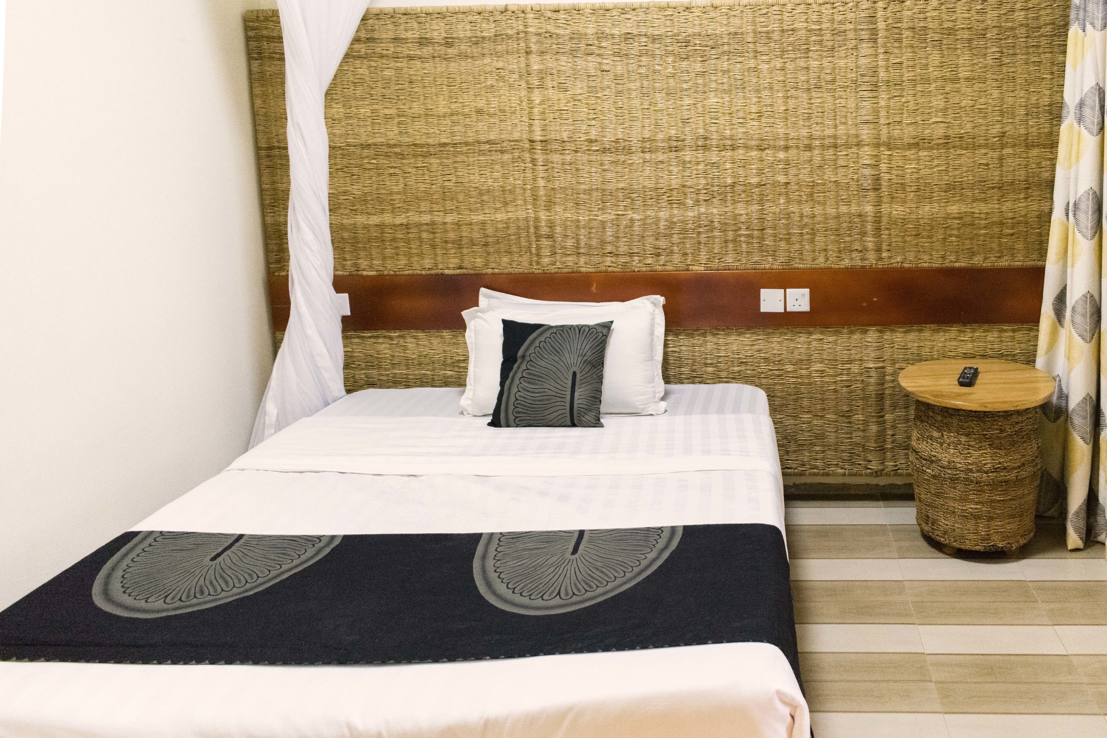 A neatly made bed with a white and black bedspread, a woven headboard, and a small round wicker side table with a remote control.
