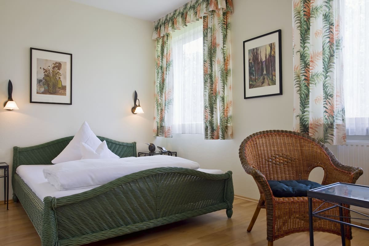 Une chambre avec un lit en osier vert, une chaise en osier, deux œuvres d’art encadrées et une fenêtre ornée de rideaux aux motifs tropicaux.
