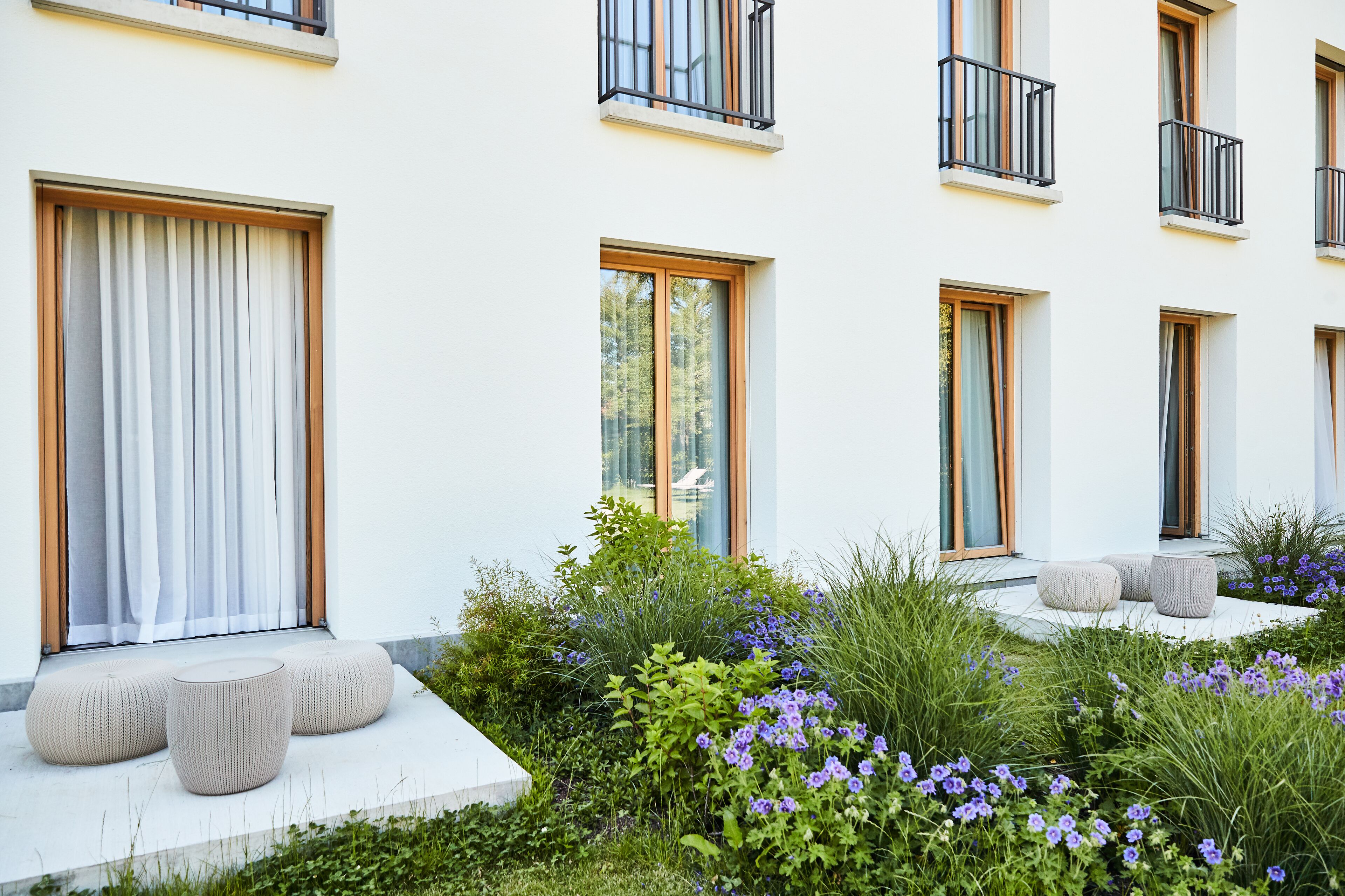 Un edificio moderno con grandes ventanales, terraza al aire libre y jardín con flores moradas.