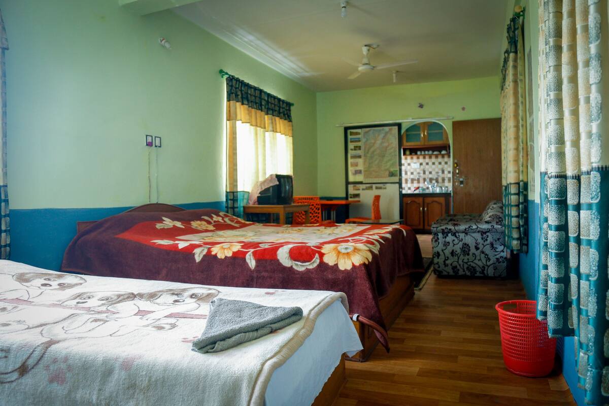 A room with two beds, a desk, a chair, a small table, a window with curtains, and a wall-mounted shelf.