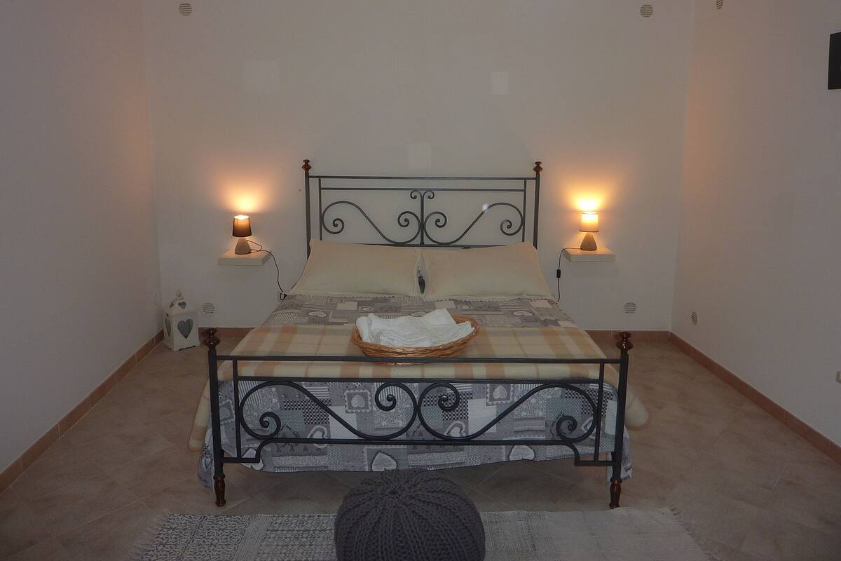 Un lit bien fait, recouvert d’une couvre-lit à motifs, avec deux lampes de chevet et un petit panier rempli de serviettes pliées posé sur le lit.