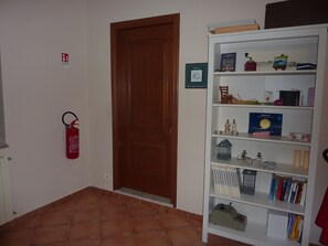 Una habitación con puerta de madera, extintor y estantería con diversos objetos, incluyendo libros, una pintura pequeña y un certificado enmarcado.