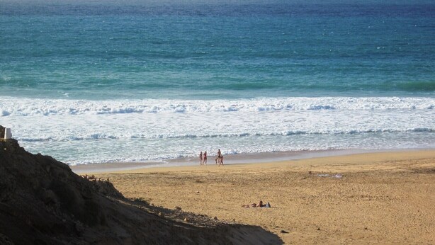 Una playa arenosa con olas rompiendo sobre la orilla, un acantilado rocoso al frente y un cielo azul despejado.