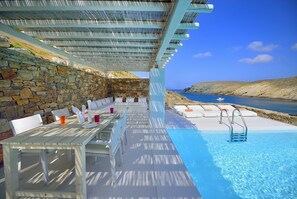 Un espace piscine extérieur agrémenté de murs en pierre, de mobilier blanc et offrant une vue dégagée sur la mer.