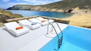 Un espace aménagé au bord de la piscine, avec des chaises longues et une vue dégagée sur une plage et un paysage rocheux.