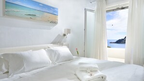 Une chambre d’hôtel avec un lit, du linge de lit blanc, un tableau représentant une plage, une table de chevet avec une lampe et une vue sur la mer par la fenêtre.