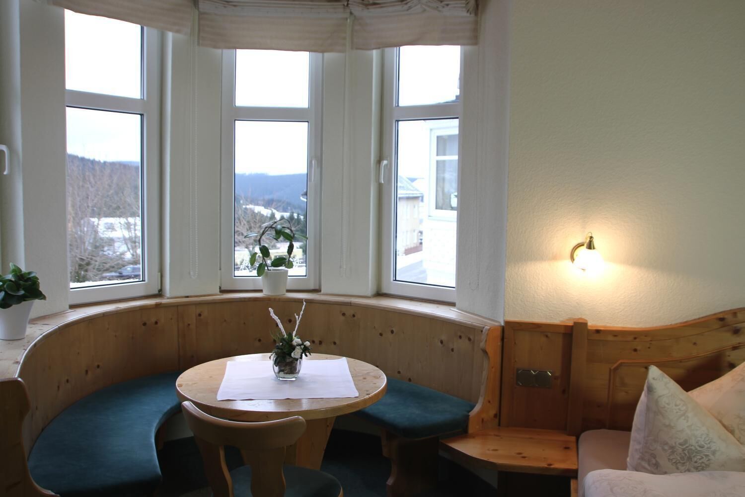 Een gezellige kamer met een zithoek met houten bank, een kleine ronde tafel met een vaas, en uitzicht op een besneeerd landschap door grote ramen.