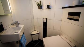 A modern bathroom with a white sink, toilet, and bidet. There are shelves with toiletries and a plant.