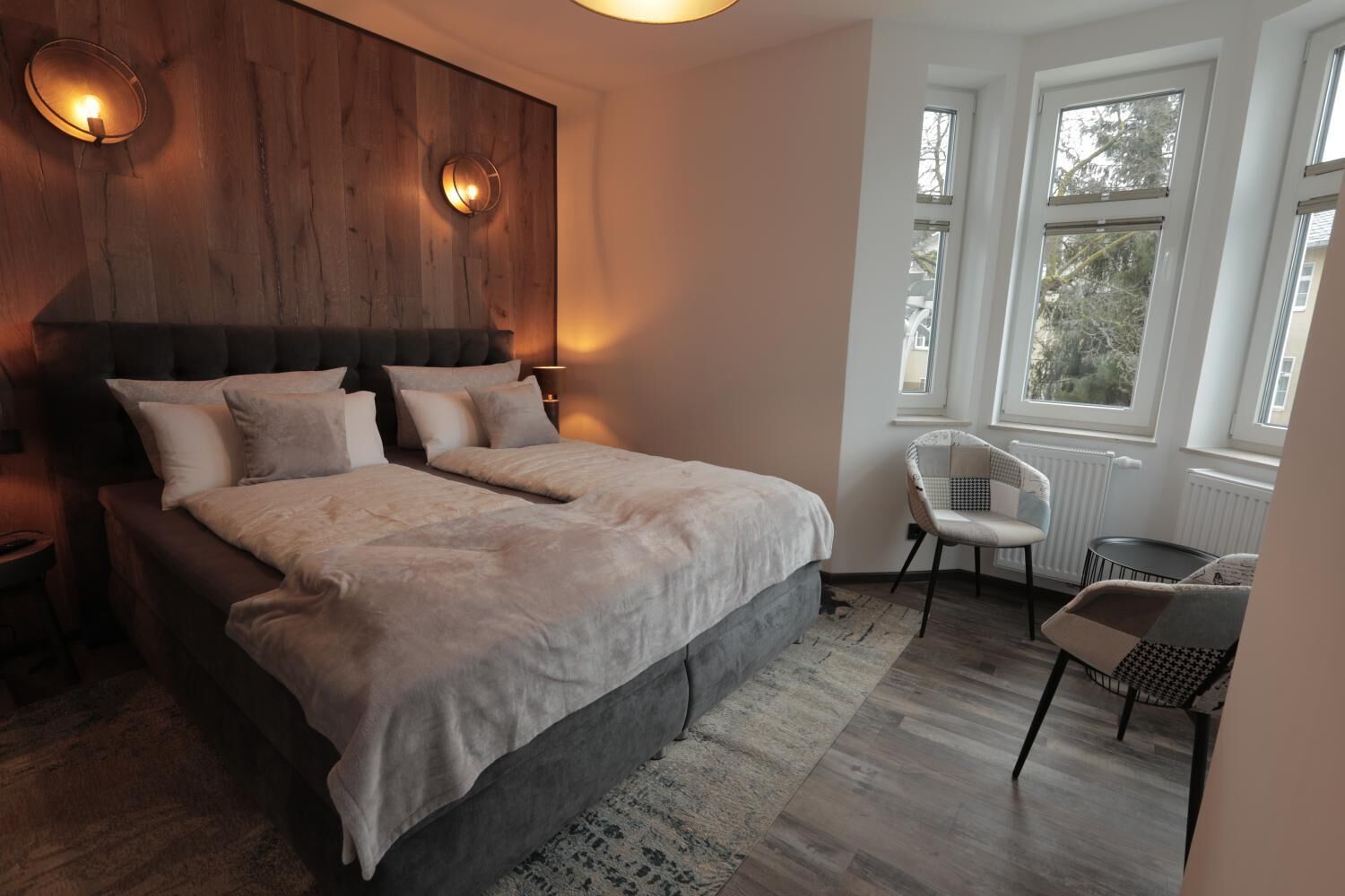 A bedroom with a large bed, two bedside lamps, a chair, and a view of trees through the window.
