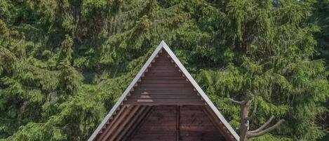 A small, triangular wooden cabin with a log bench in front, surrounded by dense forest.