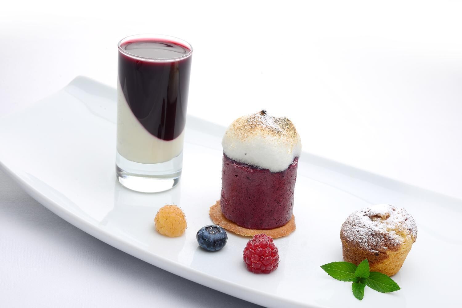 A dessert plate with a layered dessert, a small glass of red liquid, and three berries.