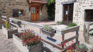 A stone building with a wooden door and window, surrounded by gravel and wooden planters with various plants. - Le Moulin de la Fortie (Viscomtat)