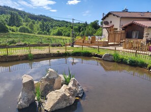 A rustic house with a stone facade and a tiled roof, surrounded by a wooden fence and a small pond with rocks and plants.
