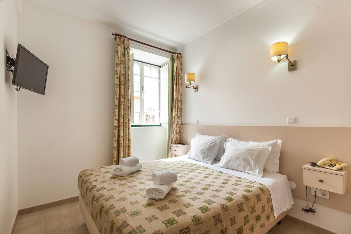 A hotel room with a bed, a bedside table with a lamp, a TV mounted on the wall, and a window with curtains.