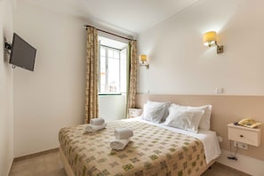 Une chambre d’hôtel avec un lit, une table de chevet avec une lampe, un téléviseur fixé au mur et une fenêtre avec des rideaux.