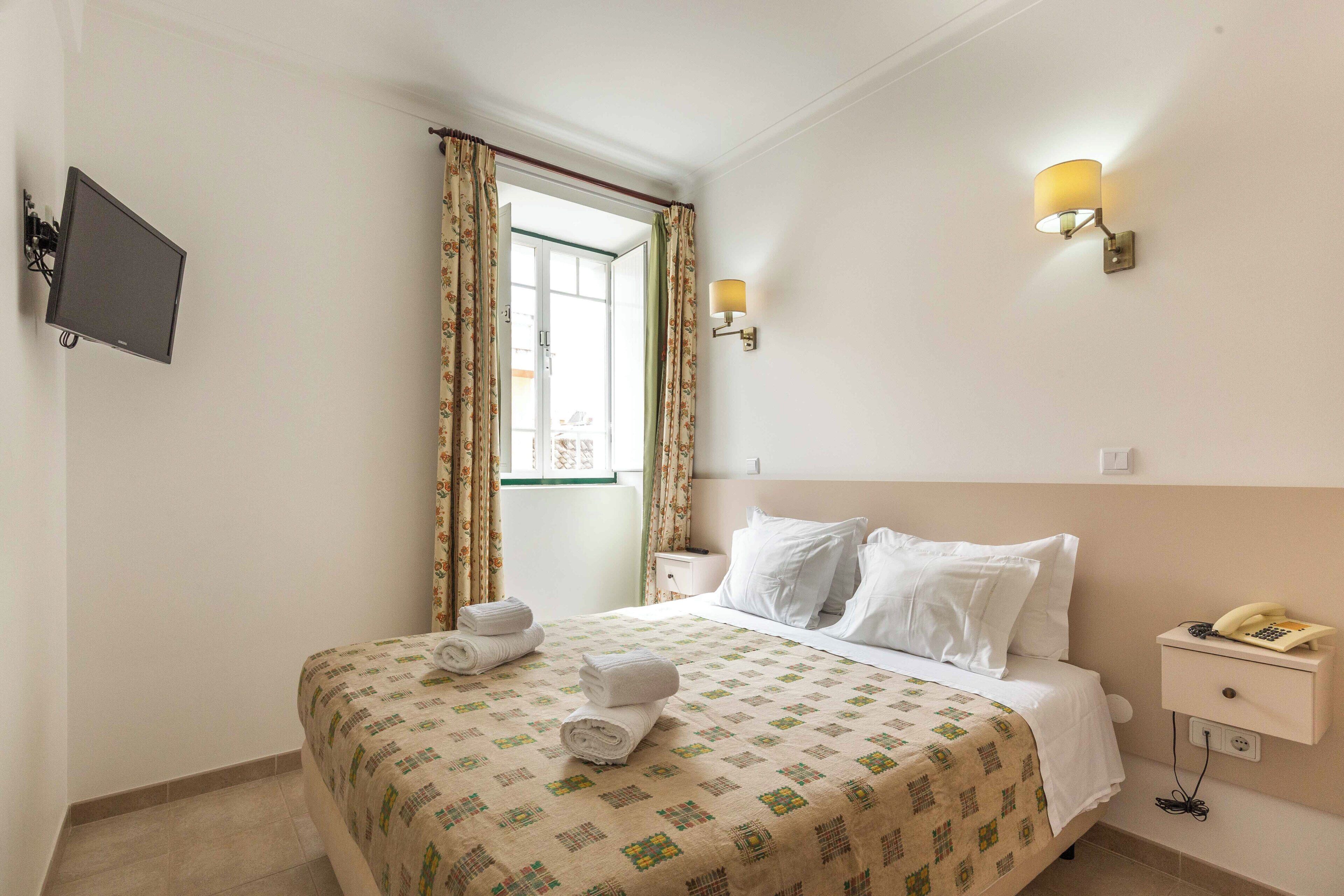 A hotel room with a bed, a bedside table with a lamp, a TV mounted on the wall, and a window with curtains.