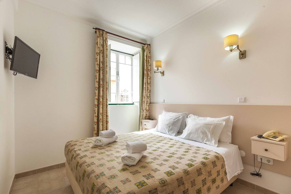 A hotel room with a bed, a bedside table with a lamp, a TV mounted on the wall, and a window with curtains.