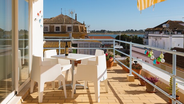 Un balcón con sillas y una mesa blancos, plantas en macetas y vistas a edificios y al mar.