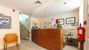 Un área de recepción con un mostrador de madera, una silla de mimbre, una escalera y varias decoraciones en las paredes, incluyendo cuadros enmarcados y un cartel de bienvenida.