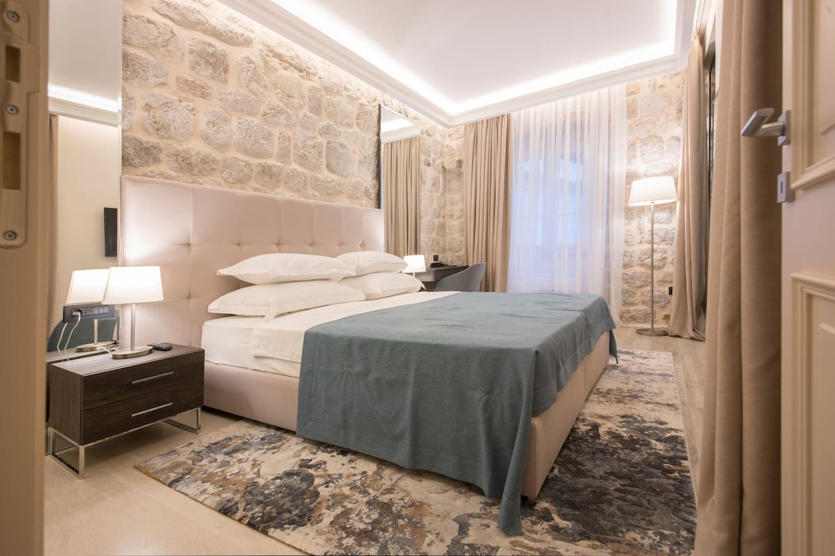 Una habitación de hotel con pared de piedra, una cama con colcha azul, una mesita de noche con lámpara y un ventanal con cortinas.