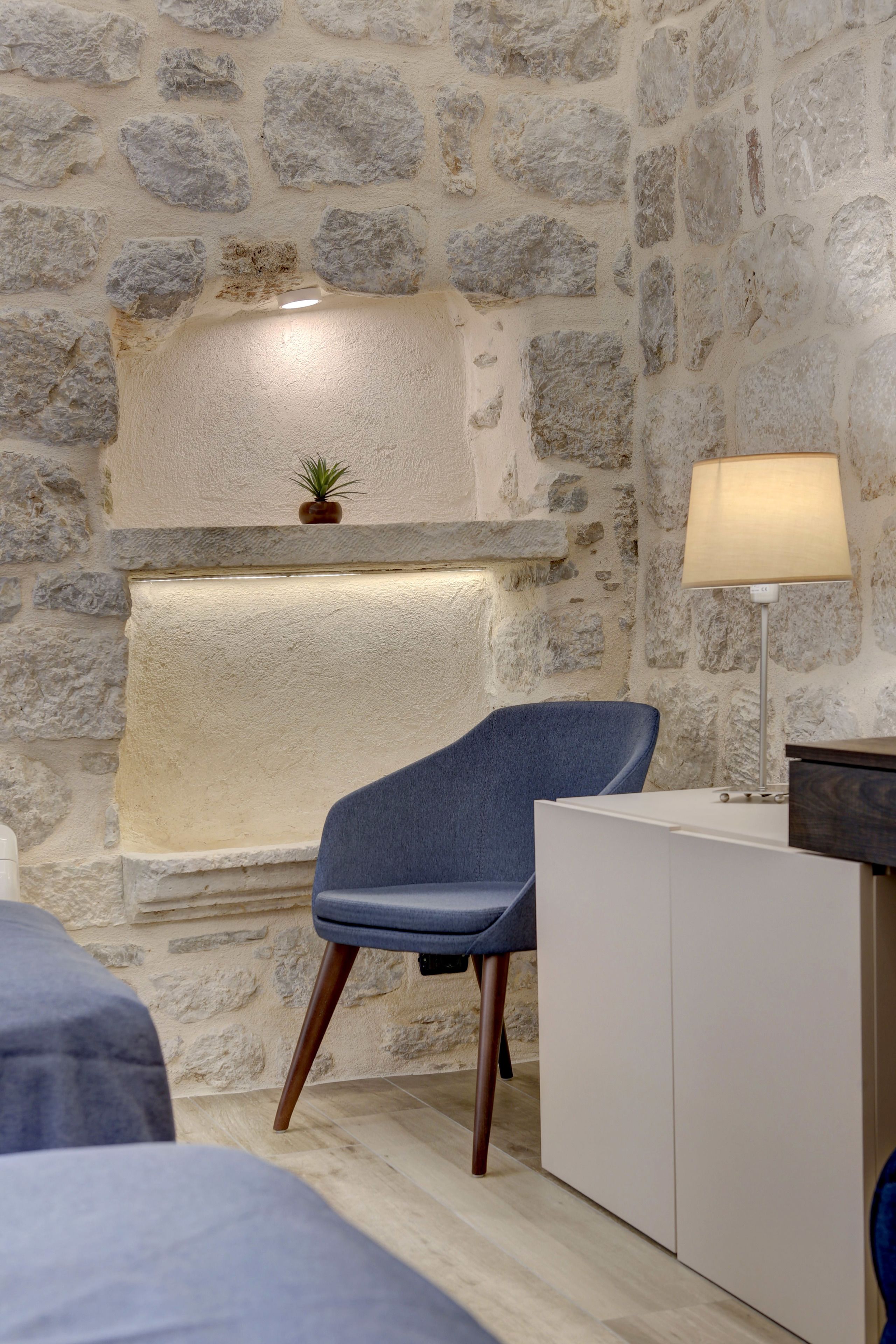 Una habitación con pared de piedra, una silla azul, una lámpara y una planta pequeña en un repisa.
