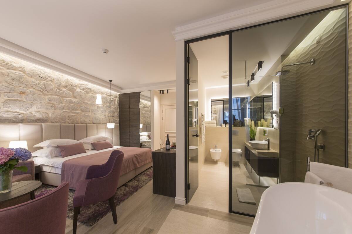 Un dormitorio moderno con una pared de acento de piedra, una cama grande, una zona de estar con una silla y vistas al baño con bañera y ducha.