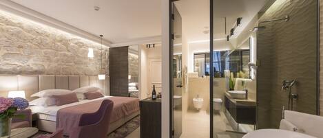 A modern bedroom with a stone accent wall, a large bed, a sitting area with a chair, and a view into the bathroom with a bathtub and shower.