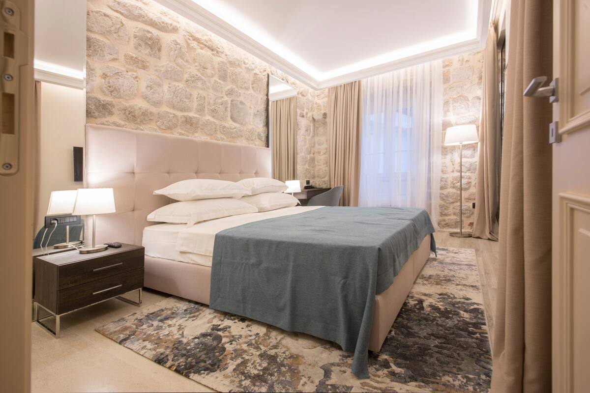 Una habitación de hotel con pared de piedra, una cama con colcha azul, una mesita de noche con lámpara y un ventanal con cortinas.