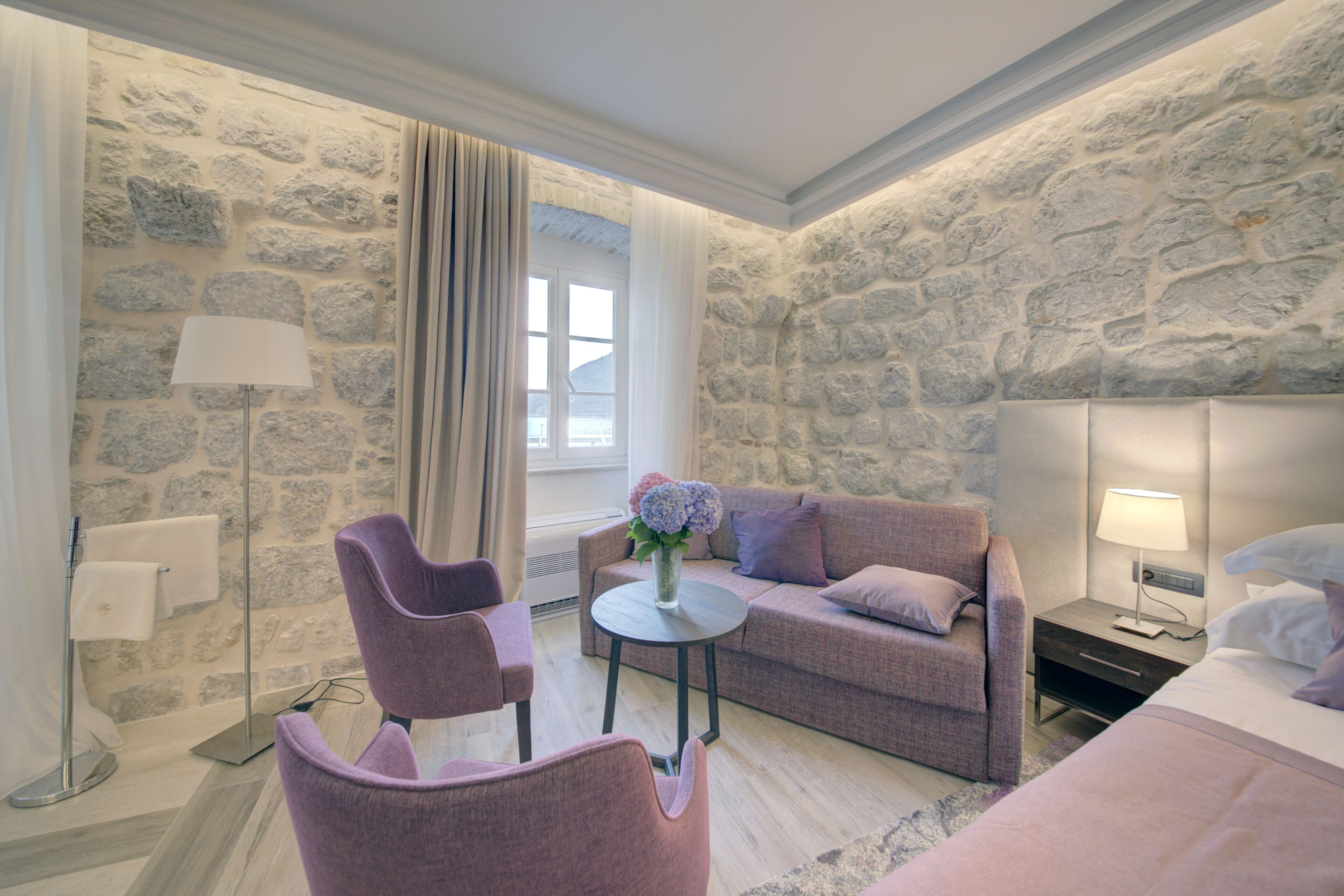 A modern hotel room with a stone wall, a sofa, a chair, a small table with a vase of flowers, and a bed with a nightstand.