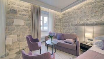 A modern hotel room with a stone wall, a sofa, a chair, a small table with a vase of flowers, and a bed with a nightstand.