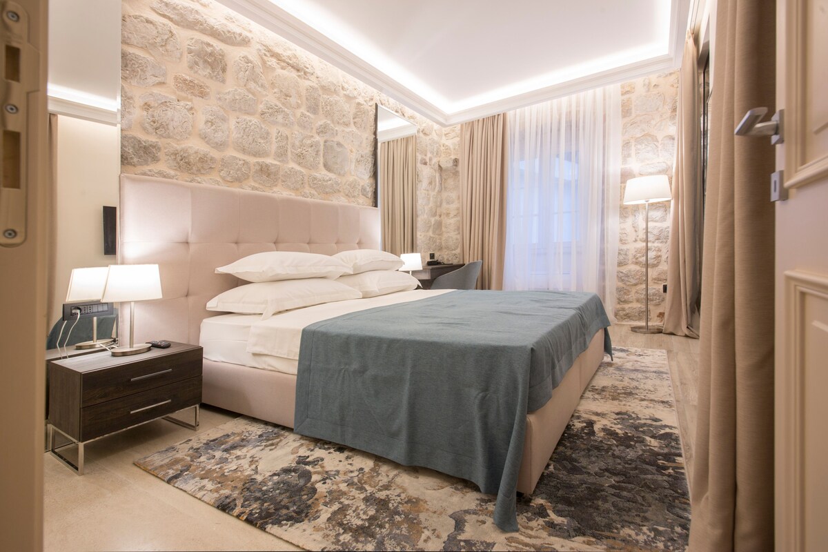 Una habitación de hotel con pared de piedra, una cama con colcha azul, una mesita de noche con lámpara y un ventanal con cortinas.