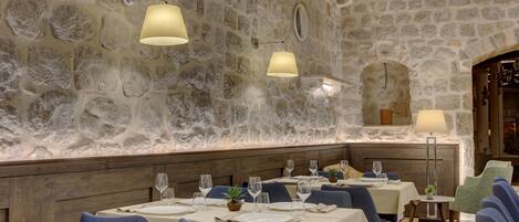 A restaurant with stone walls, blue upholstered chairs, and tables set with wine glasses and plates.