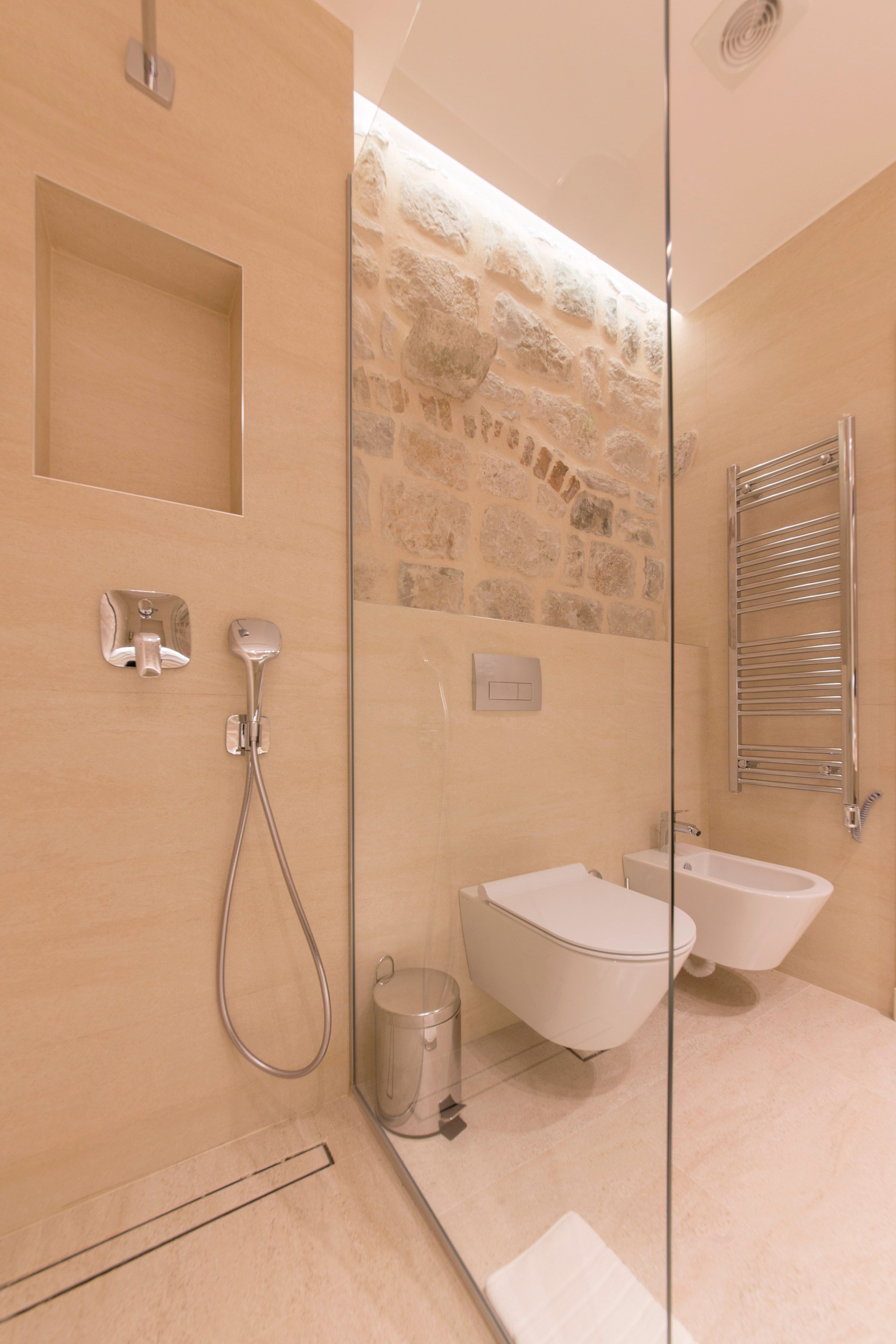 Un baño moderno con cabina de ducha de vidrio, una pared decorativa de piedra, lavabo y bidé empotrados.