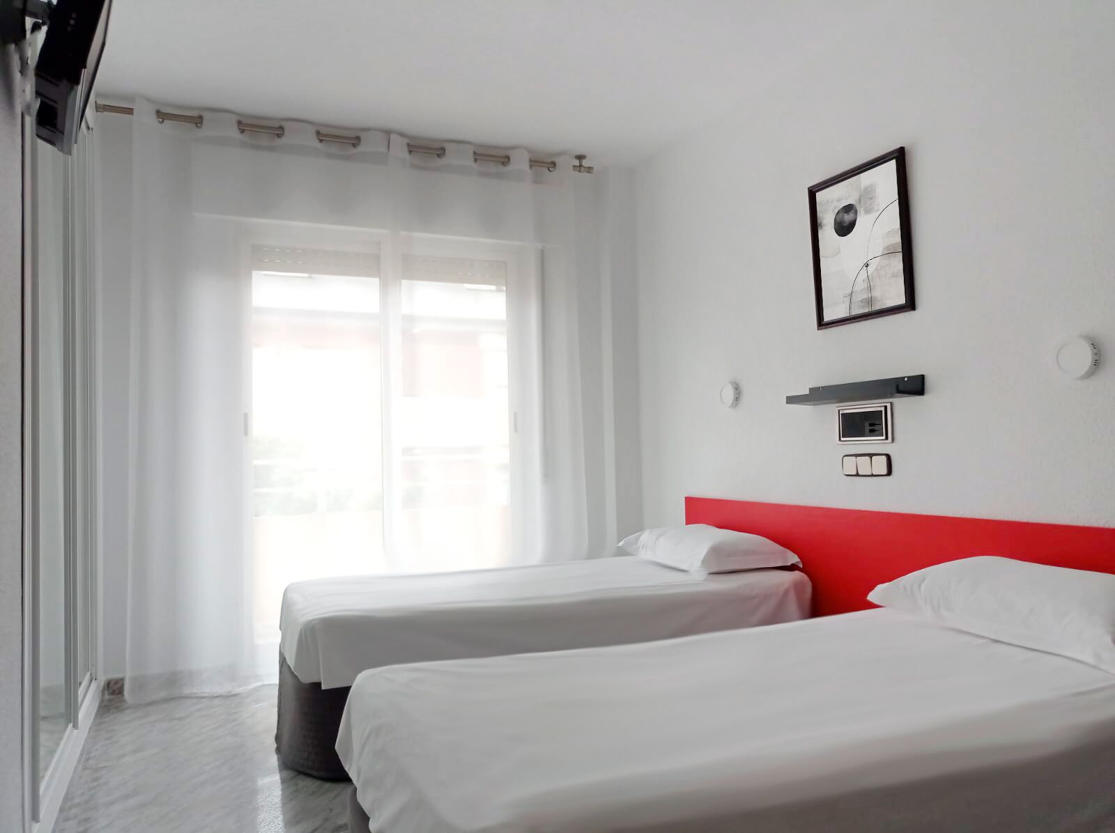 A hotel room with two beds, a red headboard, a window with sheer curtains, and a framed picture on the wall.