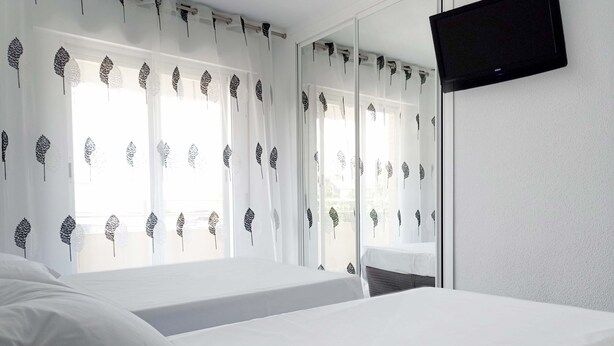 A hotel room with a bed, a TV mounted on the wall, and windows with leaf-patterned curtains. - Hostal Lady Ana Maria (Collado Villalba)