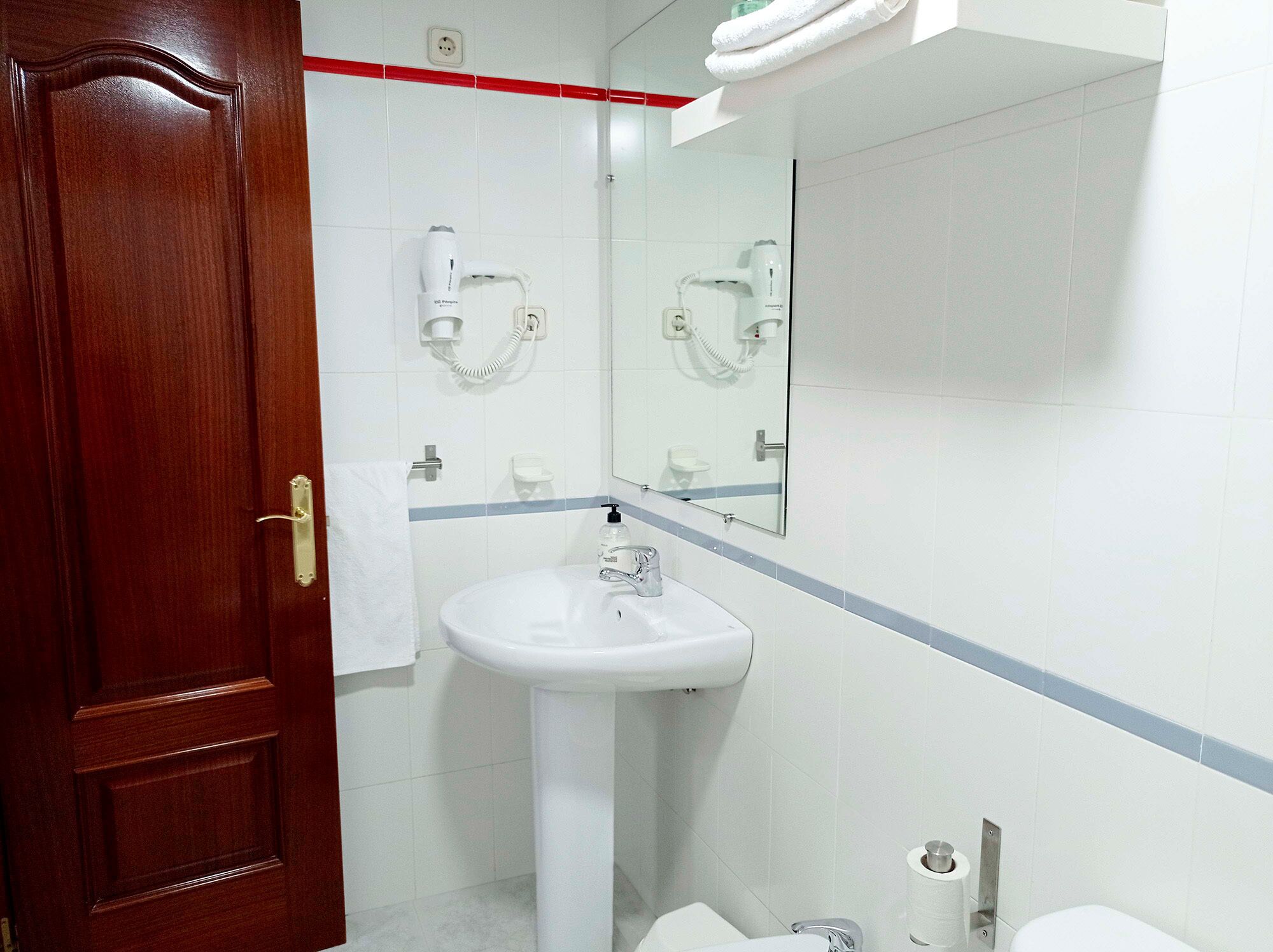 Un baño con puerta de madera, azulejos blancos, un lavamanos blanco, un espejo y un perchero para toallas.