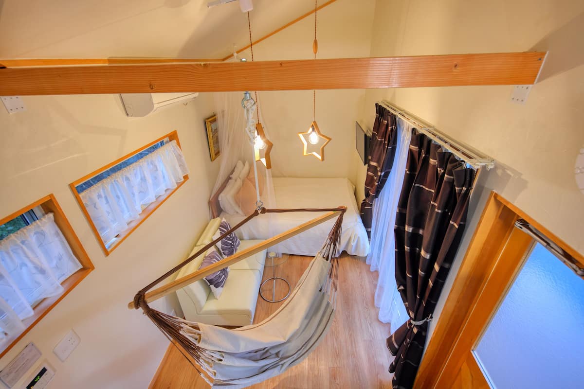 A room with a hammock bed, a wooden ceiling beam, a window with curtains, and a wall-mounted light fixture.