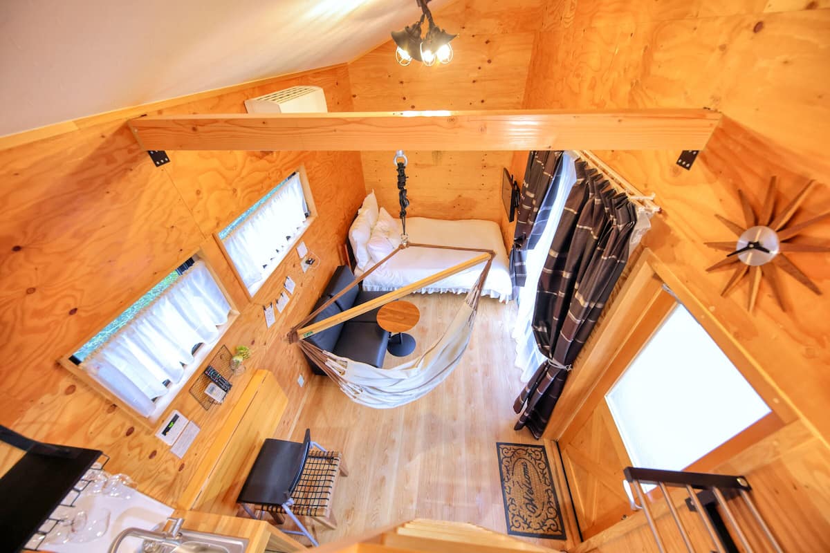A cozy cabin with a hammock, a bed, a small table, and a window.