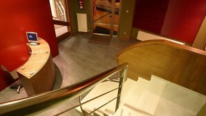 Un hall d’hôtel moderne avec un comptoir d’accueil en bois, un escalier en verre et un mur d’accent rouge.