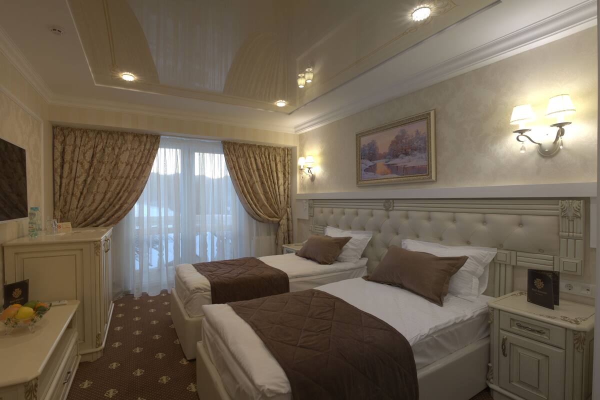 A hotel room with two beds, a large window with curtains, a painting on the wall, and a nightstand with a lamp.