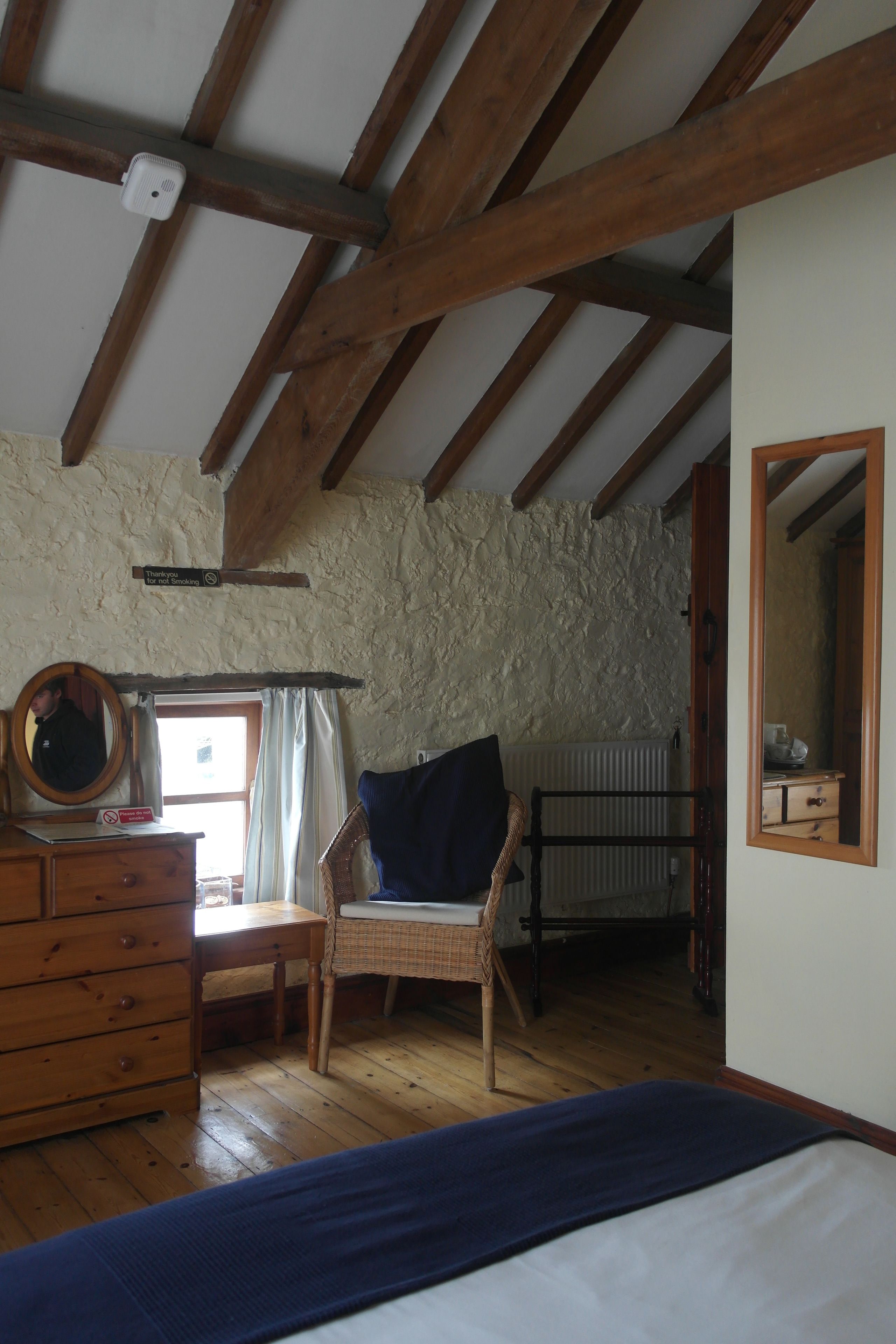 A rustic room with exposed wooden beams, a wooden dresser, a wicker chair, a bed, and a mirror.