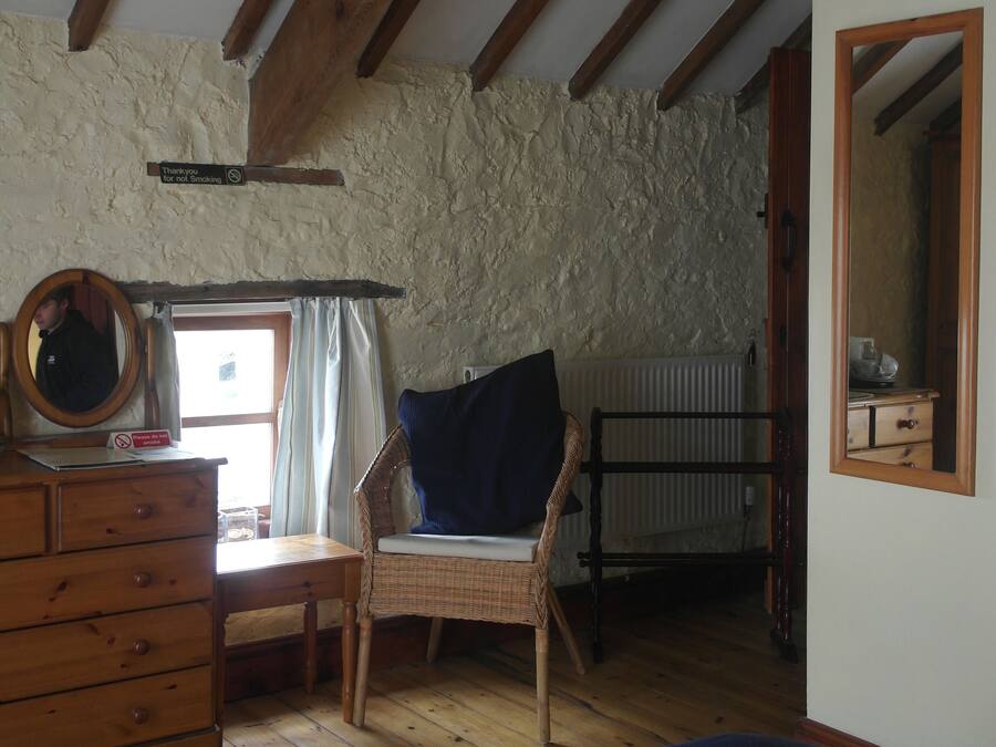 A rustic room with exposed wooden beams, a wooden dresser, a wicker chair, a bed, and a mirror.