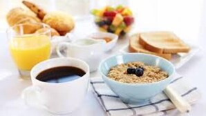 A breakfast setting with bread, fruit, a bowl of cereal, a glass of orange juice, and a cup of coffee.