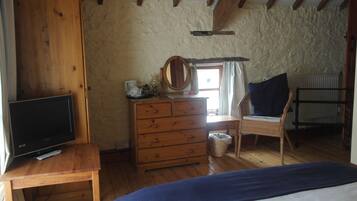 Un dormitorio con un aparador de madera, un televisor sobre una mesa de madera, una cama con manta azul, una silla de mimbre y una ventana con cortinas.