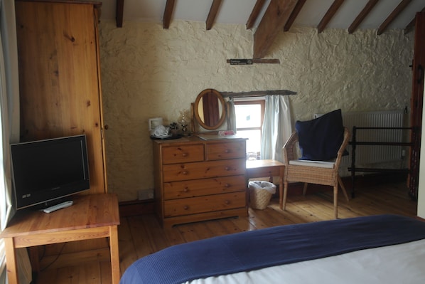 A bedroom with a wooden dresser, a television on a wooden table, a bed with a blue blanket, a wicker chair, and a window with curtains.