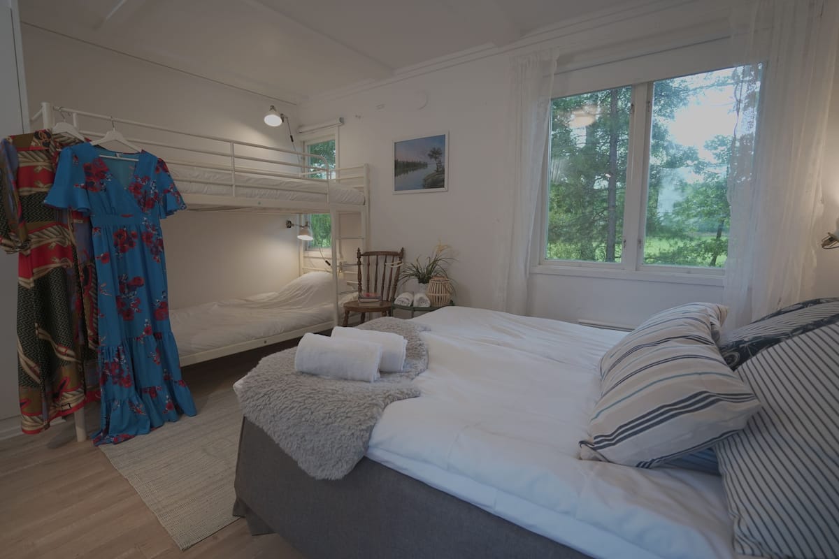 Een slaapkamer met een stapelbed, een groot raam met uitzicht op bomen, een nachtkastje met een lamp en een jurk die aan een rek hangt.