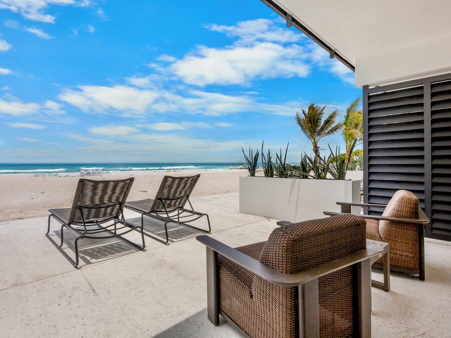 Un patio con dos sillones de mimbre y una mesa, con vista a una playa con palmeras y un cielo despejado.